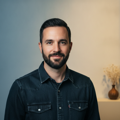 Portrait of James Henderson, a man with short dark hair and a beard, smiling confidently in a dark denim shirt against a crea